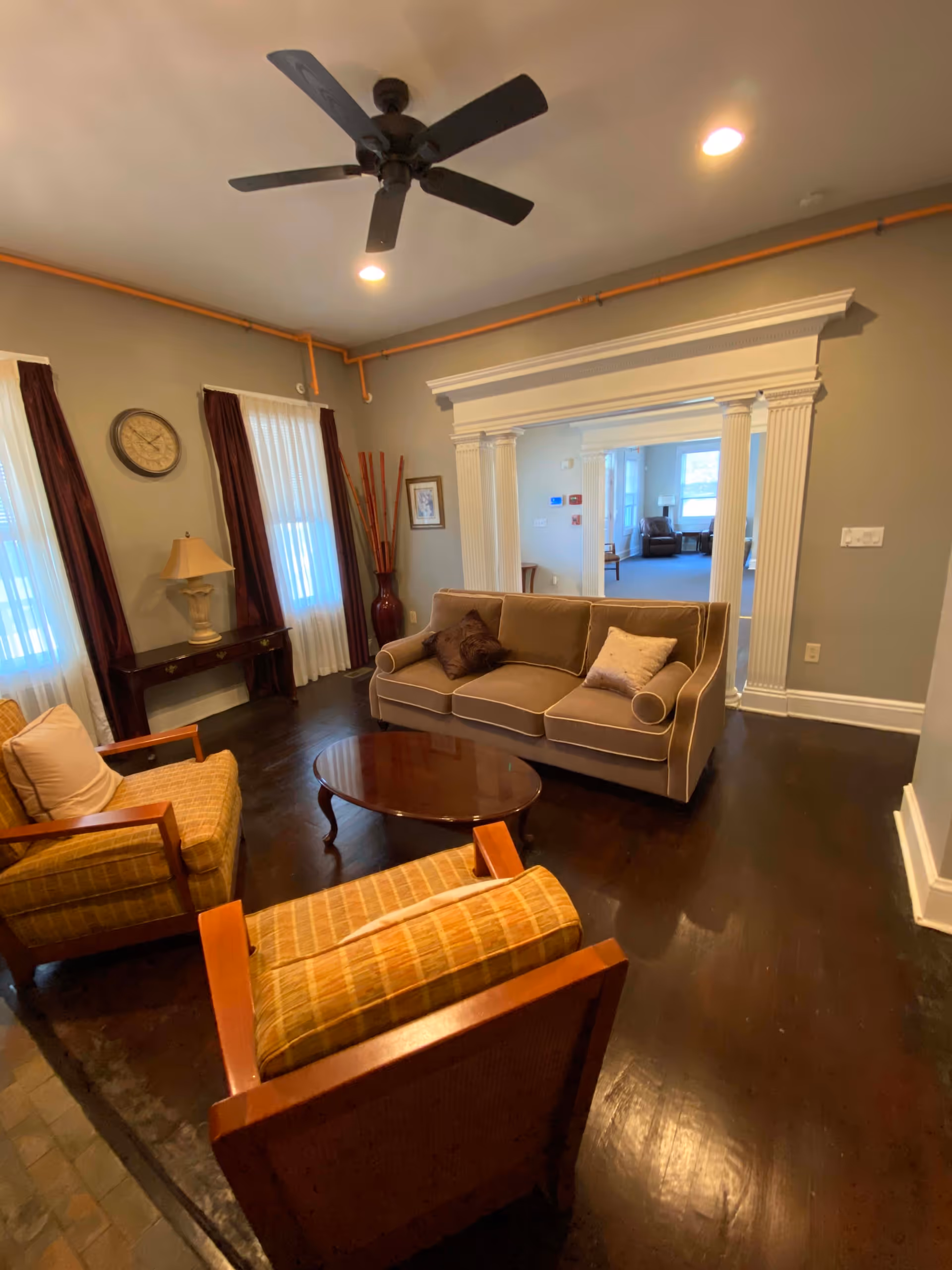 A cozy living room with a brown sofa, two cushioned armchairs, a wooden coffee table, and a side table with a lamp. The room features dark hardwood floors, beige walls, two windows with white curtains and dark drapes, a ceiling fan, and an ornate white doorway leading to another room with additional seating.