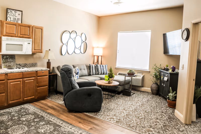A cozy living room area with a gray sectional sofa adorned with patterned pillows, a dark gray recliner chair, and a round wooden coffee table. The room features a wall-mounted TV, a black cabinet with plants on top, and a window with closed blinds. To the left, there is a small kitchen area with wooden cabinets, a microwave, and a countertop. The floor has a mix of wood and patterned carpet, and the walls are painted beige with decorative mirrors and framed signs.