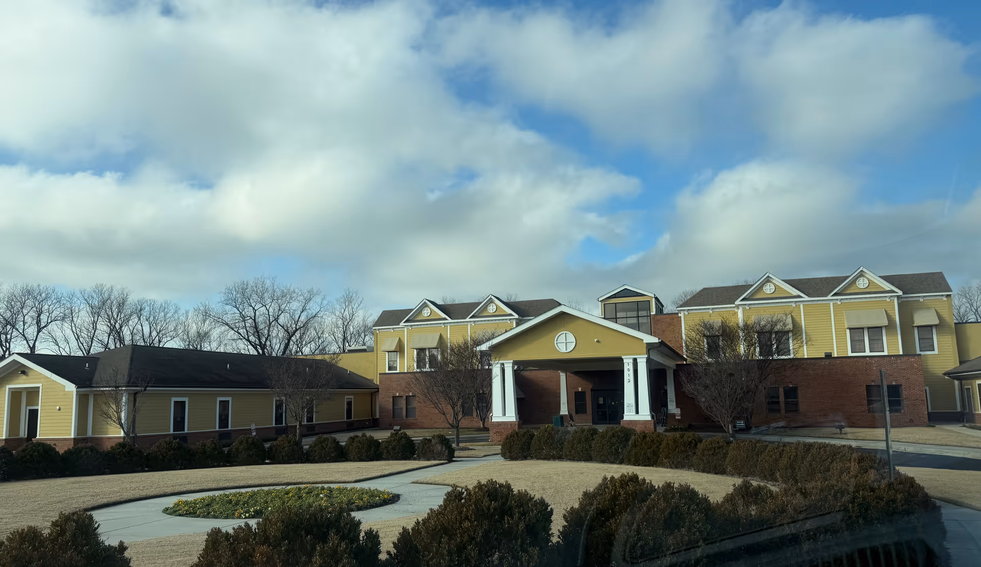 Front exterior of a yellow-and-brick senior living facility with a covered entrance and circular driveway under a partly cloudy sky.