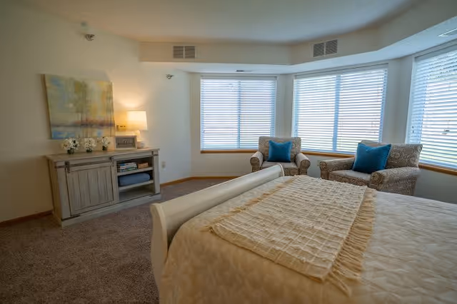 A bright bedroom with a large bed covered in a beige quilt and a textured throw. Two patterned armchairs with blue cushions are positioned near a bay window with white blinds. A wooden cabinet with decorative items and a lamp is against the wall, and a painting hangs above it.
