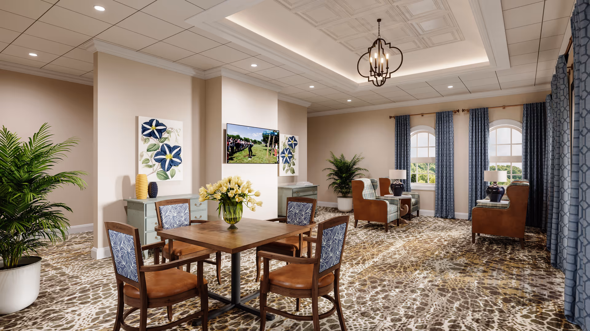 A bright and spacious common area with a wooden table and four chairs in the foreground, decorated with a vase of yellow flowers. The room features patterned carpet, beige walls, two large potted plants, and three arched windows with blue curtains letting in natural light. There are two armchairs with side tables and lamps near the windows, a wall-mounted TV, and floral artwork on the walls.
