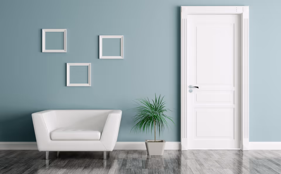 Minimalist room with a white armchair, potted plant, three empty square frames on a teal wall, and a closed white door.