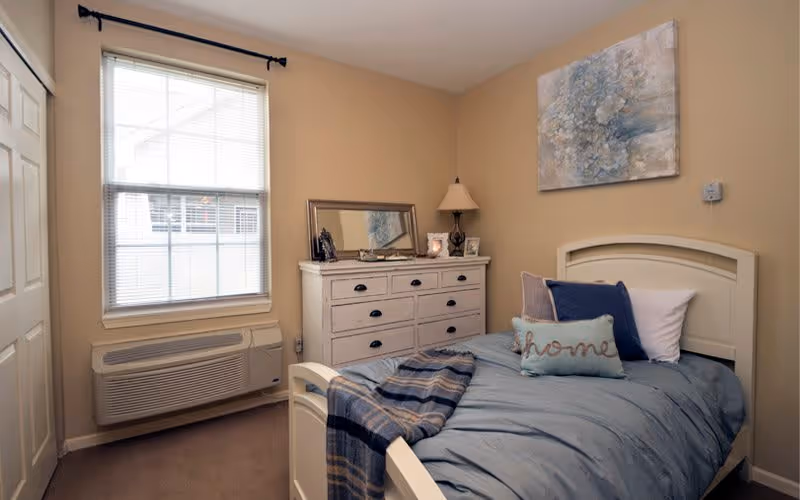 A cozy single bedroom with a white bed dressed in blue bedding, a white dresser topped with a mirror and lamp, and a window with blinds.