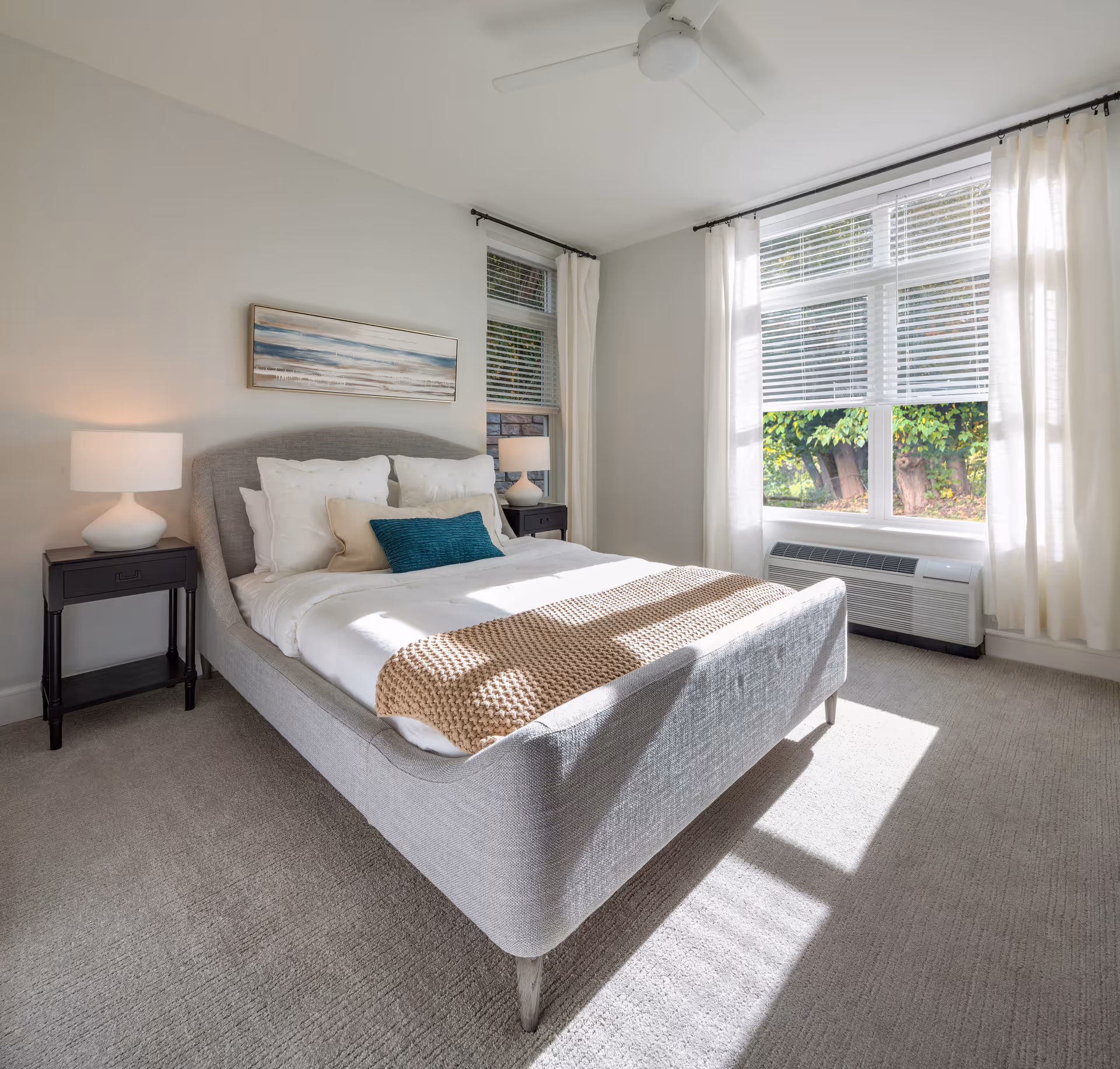 A bright and airy bedroom with a large window letting in natural light. The room features a gray upholstered bed with white bedding, multiple pillows including a teal accent pillow, and a beige knitted throw blanket. There are two black nightstands on either side of the bed, each with a white lamp. A ceiling fan is mounted above, and a landscape painting hangs on the wall above the bed.