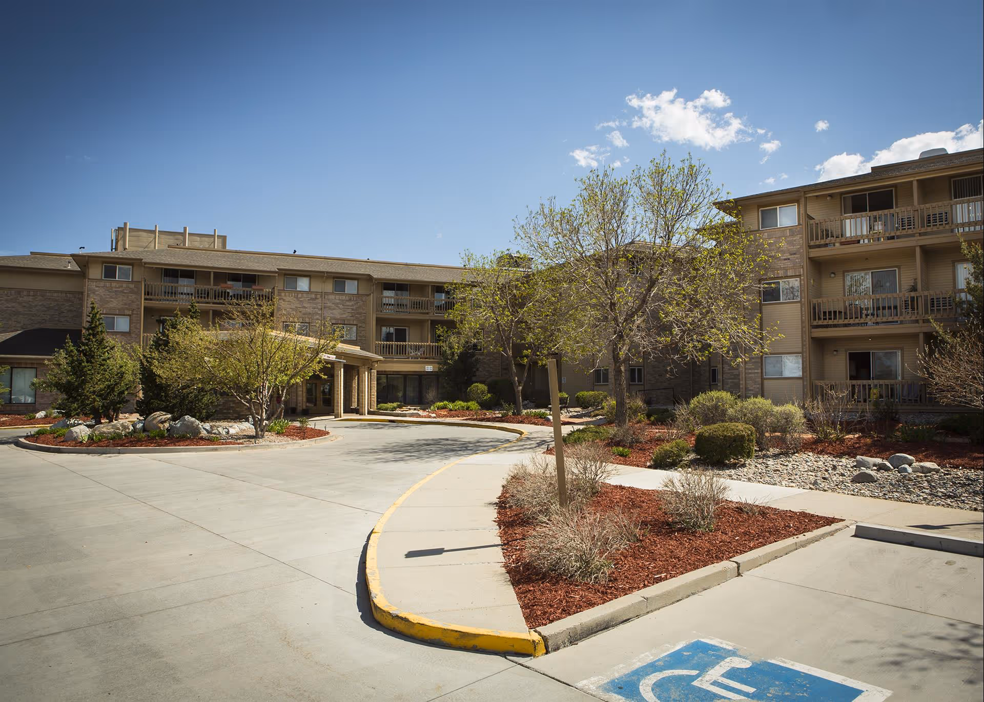 Front entrance and driveway of a multi-story senior living facility with landscaped beds, trees, balconies, and a blue sky.