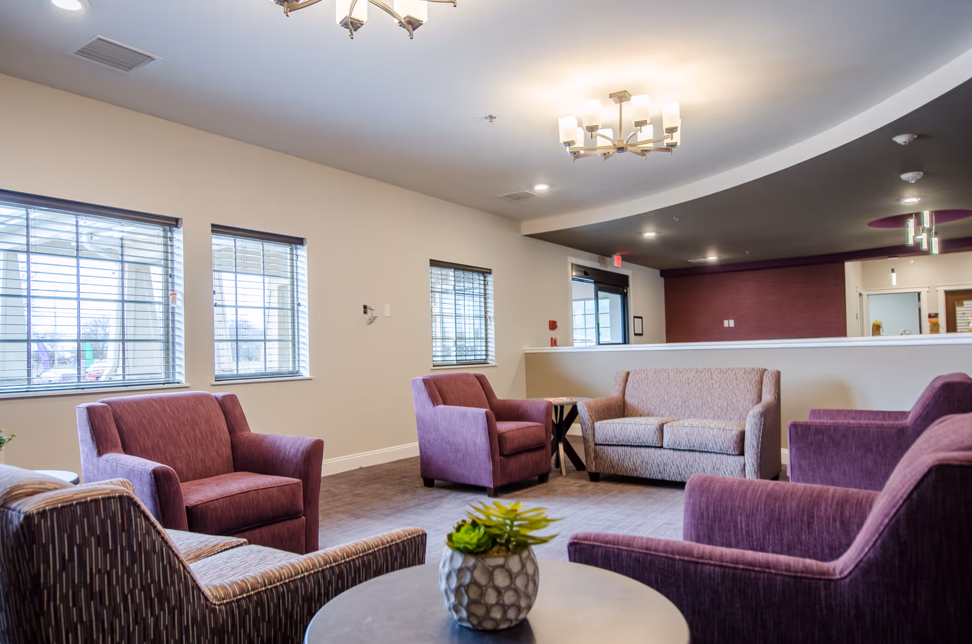 Communal lounge with purple armchairs, a loveseat, and a small table with a plant under ceiling lights.