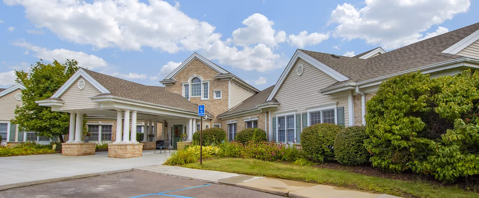 Exterior view of a senior living facility with beige siding and brick accents, featuring a covered entrance supported by white columns, surrounded by green bushes and trees under a partly cloudy sky.