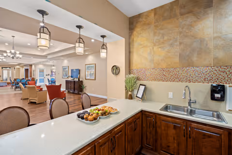Interior view of a senior living facility showing a kitchen counter with a sink, a bowl of fruit, and a plate of pastries. The counter overlooks a spacious common area with chairs, tables, and a television. The walls have warm tones with tile backsplash in the kitchen area and pendant lights hanging from the ceiling.