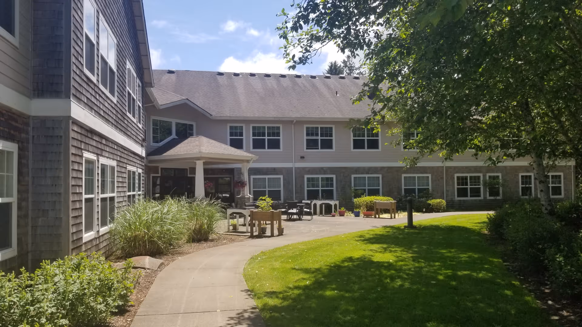 Outdoor view of a senior living facility with a paved walkway curving through a green lawn and landscaped bushes. The building has two stories with multiple windows and a covered entrance. There are tables and chairs placed on the patio area near the entrance. Trees provide shade on the right side of the image under a partly cloudy sky.