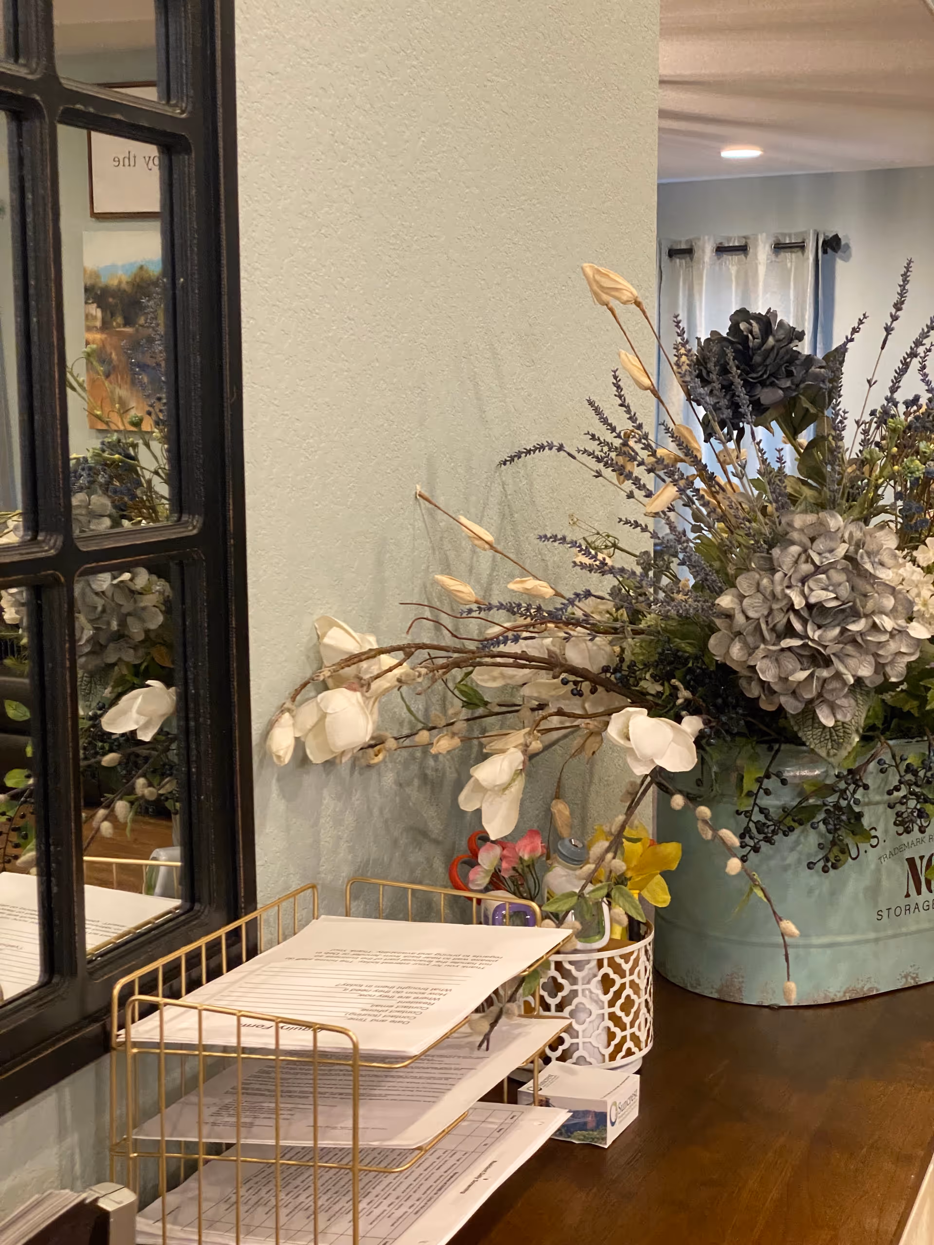 A decorative arrangement on a wooden surface featuring a large metal container filled with artificial flowers including white magnolias, lavender, and hydrangeas. Next to it is a gold wire paper organizer holding documents, a small patterned container with pens, and a box of tissues. A black-framed mirror is mounted on the light-colored wall behind the arrangement, reflecting part of the room.