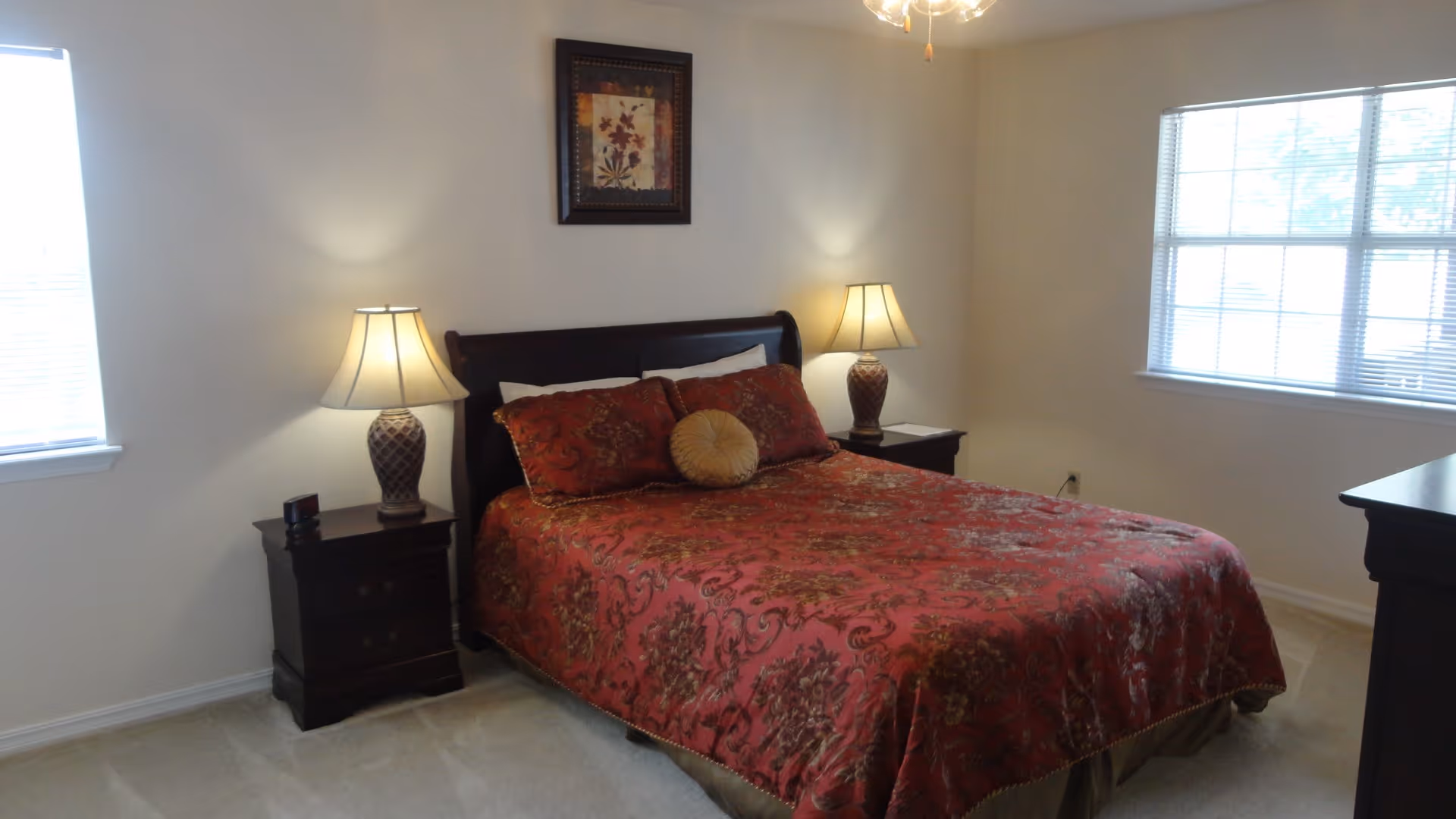 A bedroom with a double bed covered in a red patterned bedspread, two matching pillows, and a round decorative pillow. There are two dark wooden nightstands on either side of the bed, each with a lamp. The room has beige walls, two windows with blinds, and a framed floral artwork above the bed.