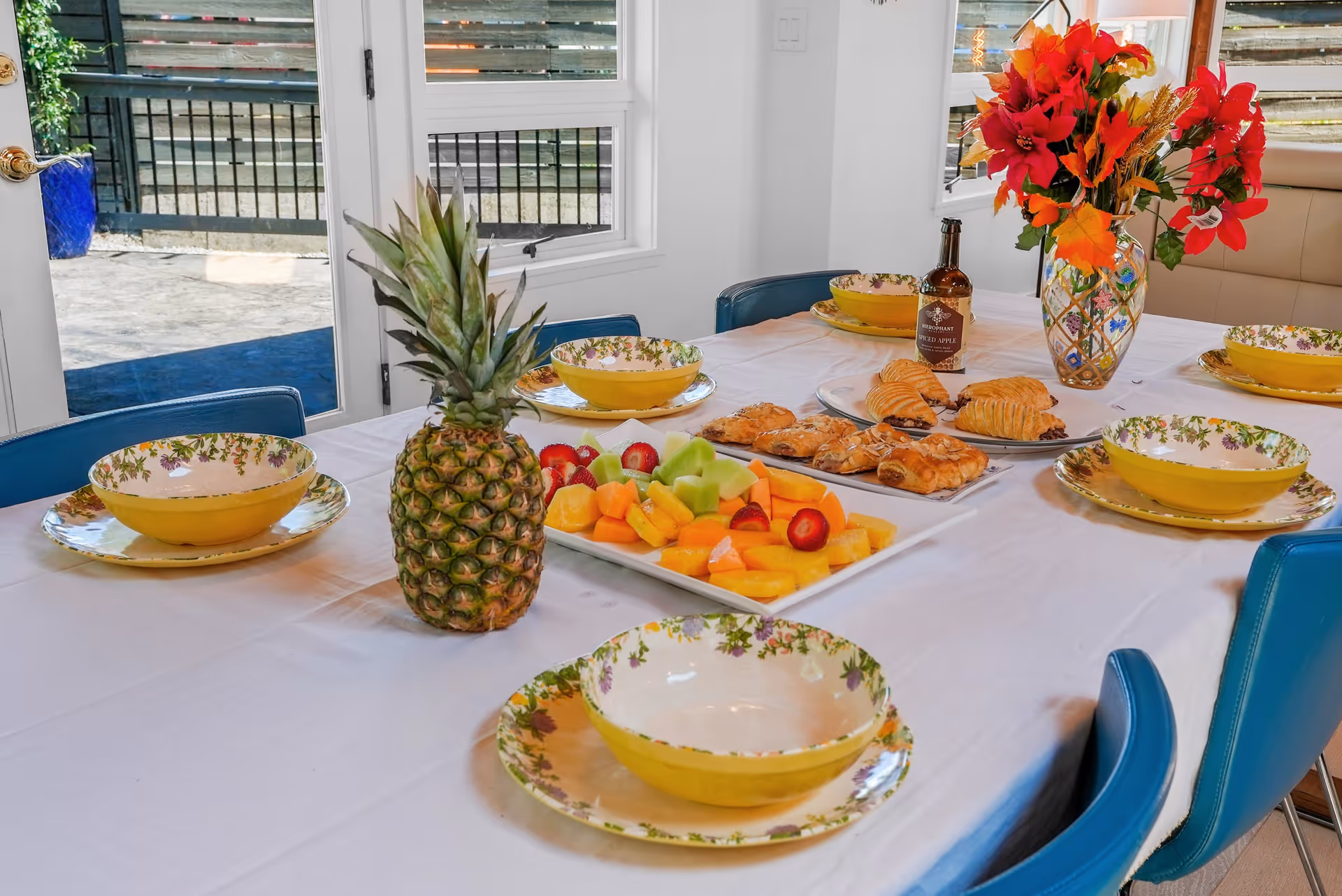 A dining table set with floral-patterned yellow bowls and plates, a white tablecloth, a centerpiece vase with red and orange flowers, a plate of assorted fresh fruit including pineapple, melon, and strawberries, and a plate of pastries. The room has large windows and a door leading to an outdoor patio area.