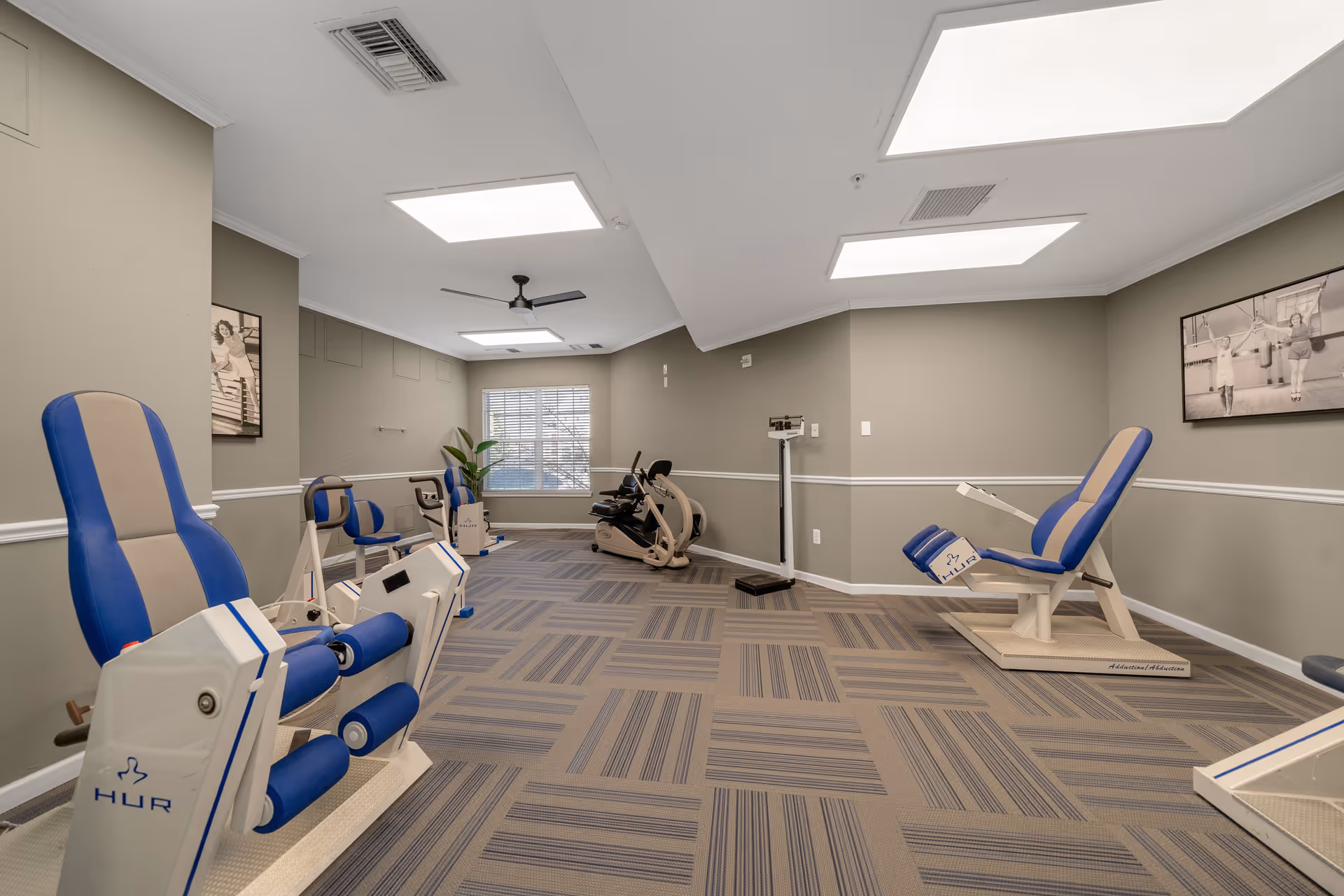 A well-lit exercise room with various fitness equipment including leg press machines and a stationary bike. The room has beige walls with white trim, a patterned carpet, ceiling lights, a ceiling fan, and a window with blinds. Black and white framed photos hang on the walls.