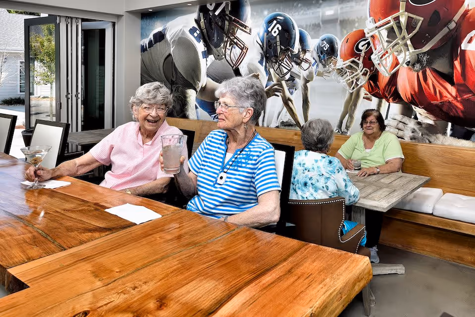 Four elderly women sitting at wooden tables inside a room with large windows and a mural of football players on the wall. Two women are seated at a bar-style table holding drinks and smiling, while the other two women are seated at a booth table engaged in conversation.