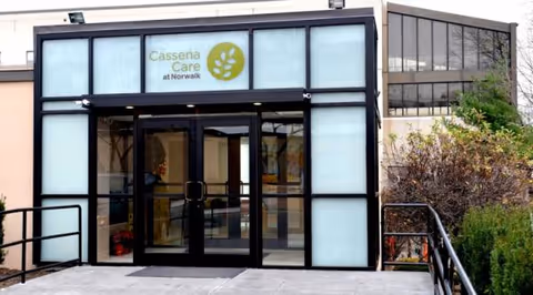 Glass-front entrance of Cassena Care at Norwalk with automatic doors and surrounding shrubs.
