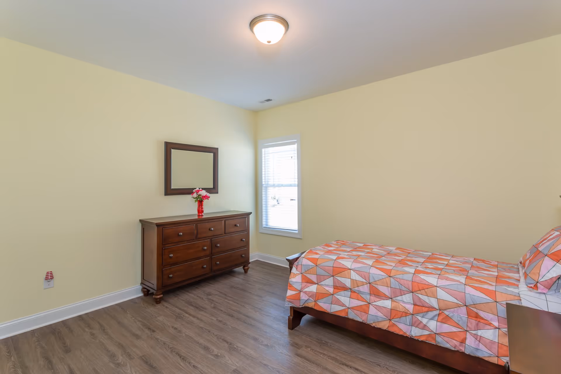 A simple bedroom with light yellow walls, a wooden dresser with a mirror above it, a vase with flowers on the dresser, a window with white blinds, and a bed with a colorful geometric patterned bedspread.