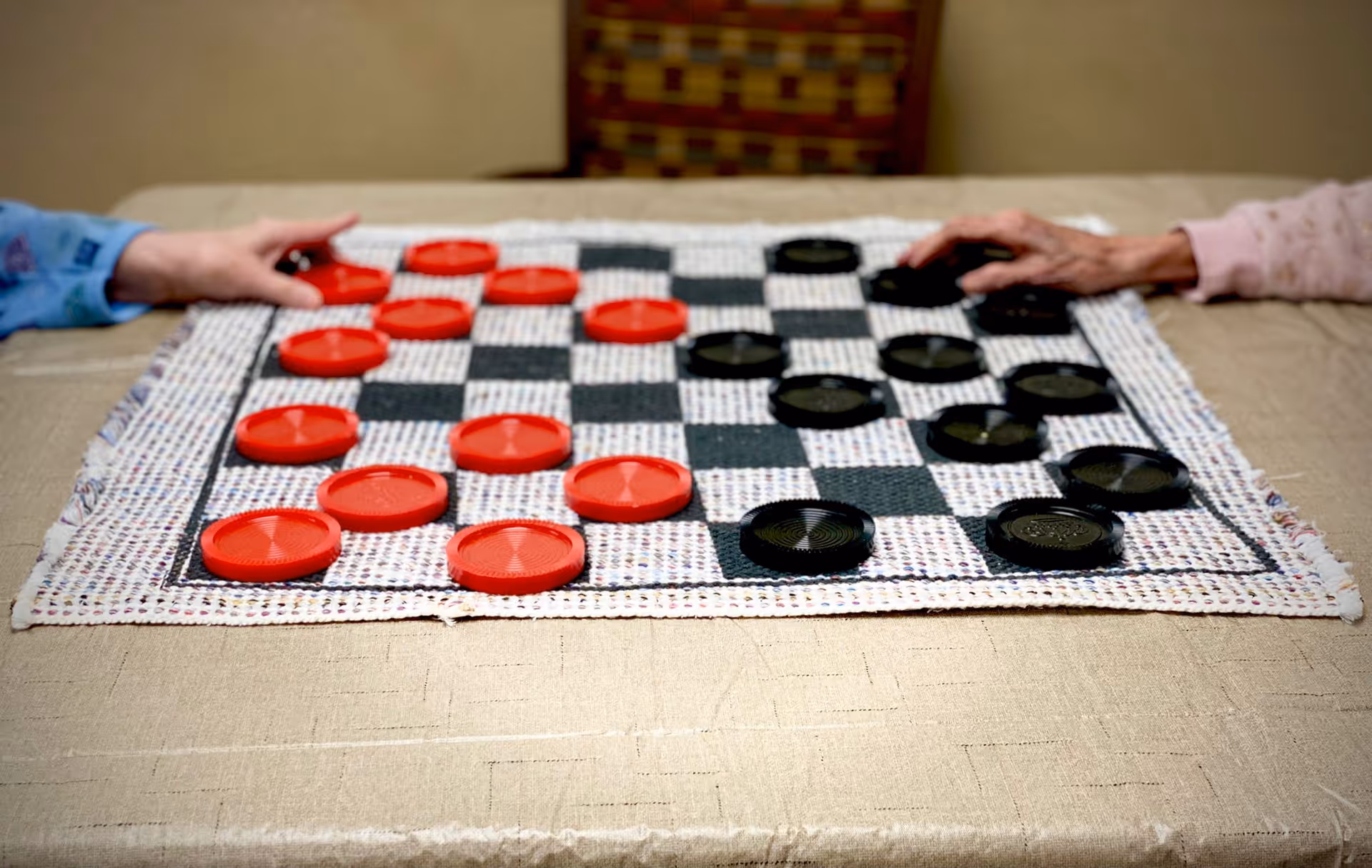 Two elderly individuals playing a game of checkers on a checkered board with red and black pieces on a beige tablecloth.