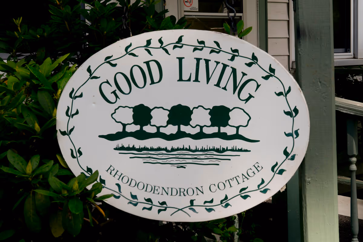 An oval white sign with green text and graphics hanging outside a building. The sign reads 'GOOD LIVING' at the top and 'RHODODENDRON COTTAGE' at the bottom, with a graphic of five trees and a body of water in the center. The sign is surrounded by green leafy plants and is attached to a green post near a porch with stairs.