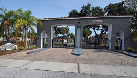 Entrance gate to The Villas of Casa Celeste, featuring a gated archway with a tiled roof, palm trees, and a sign displaying the facility's name on the left side.