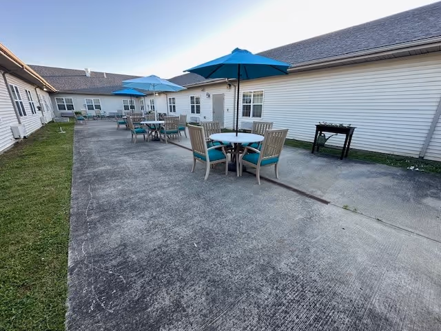 Outdoor patio area at The Gardens of Green Ridge with several round tables and chairs, each table shaded by a blue umbrella. The patio is surrounded by a single-story building with white siding and multiple windows. There is a small grill or barbecue on the right side near the building.