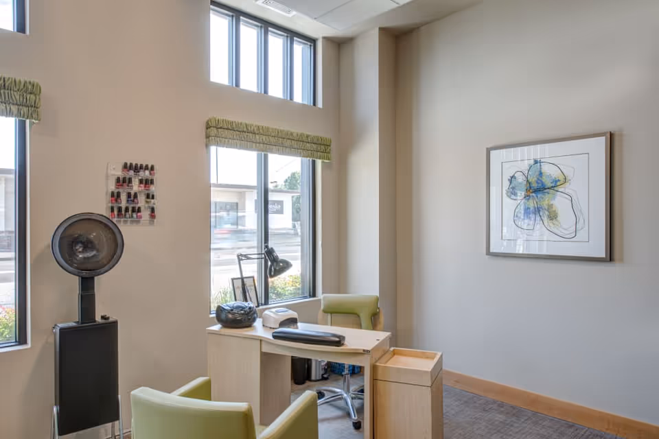 A small salon area with a manicure table and two green chairs. On the table are manicure tools and a lamp. A wall-mounted nail polish rack with various colors is visible next to a large hair dryer. The room has tall windows with green valances and a framed abstract artwork on the wall.