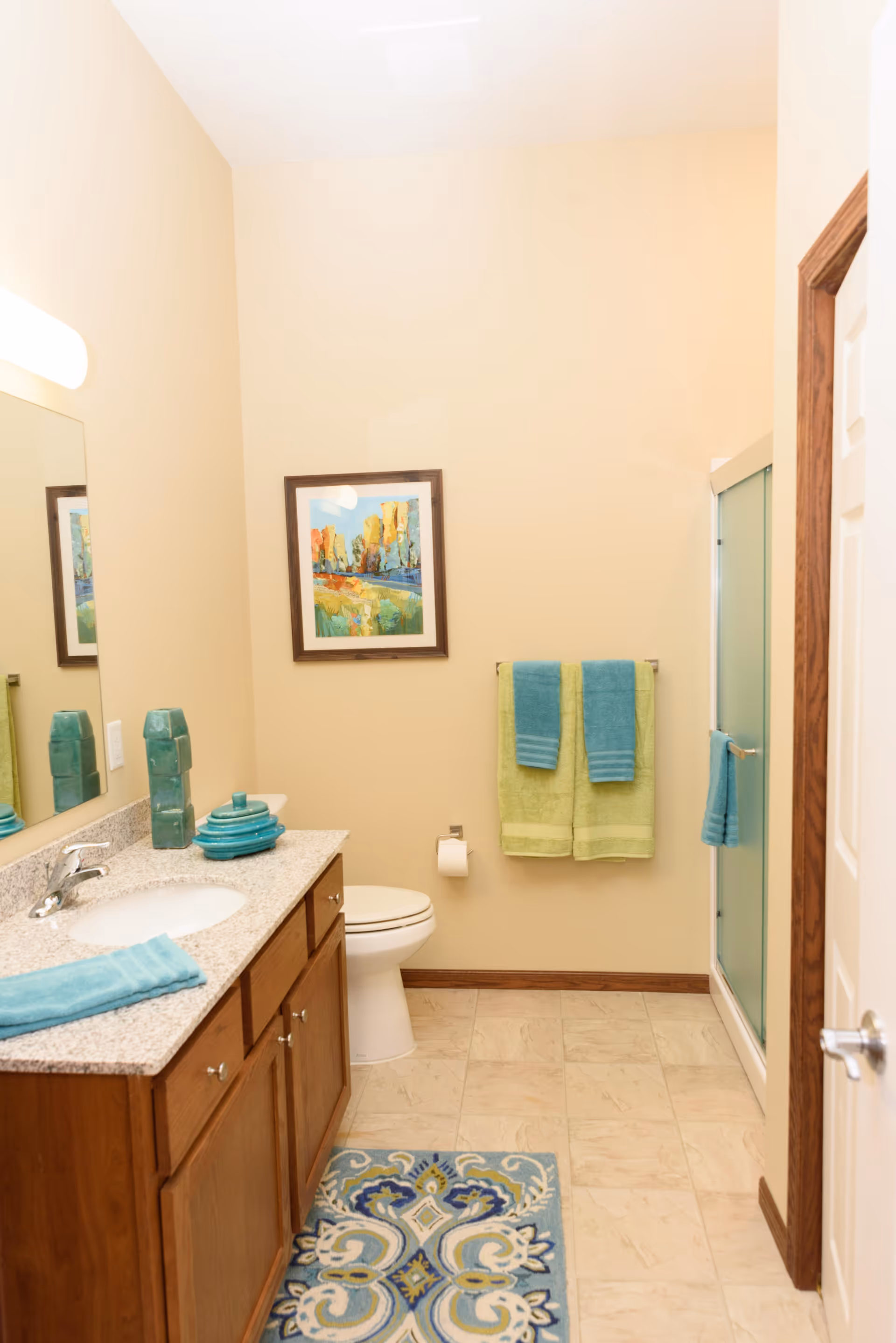 A clean bathroom with beige walls and tiled floor. There is a wooden vanity with a granite countertop, a sink, and turquoise decorative items and towels. A toilet is positioned against the far wall beneath a colorful framed painting. Two green and blue towels hang on a towel rack next to a glass-enclosed shower. A patterned blue and beige rug is on the floor in front of the vanity.