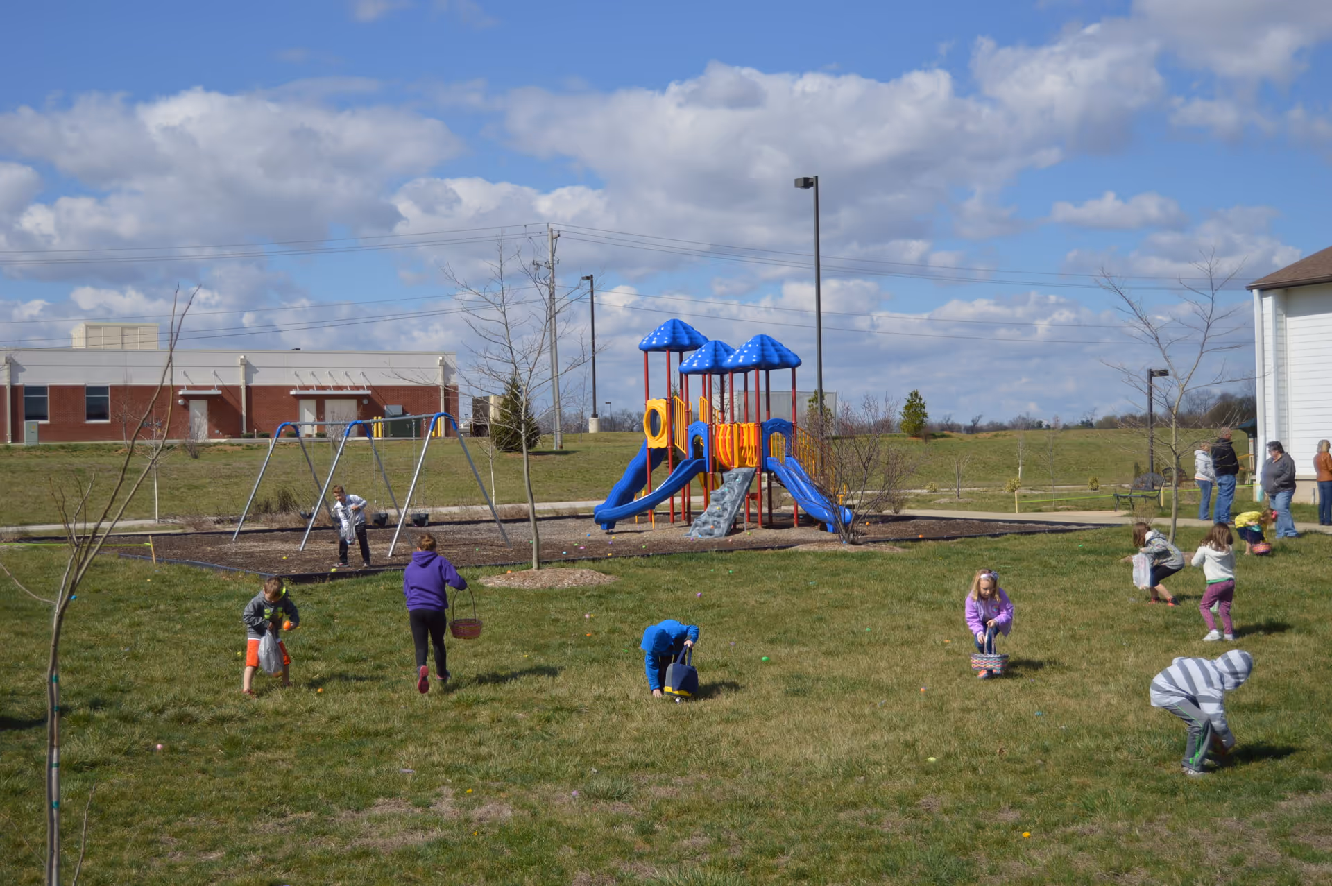 Children playing and gathering Easter eggs on a grassy playground area with a blue and orange play structure and swings in the background under a partly cloudy sky.