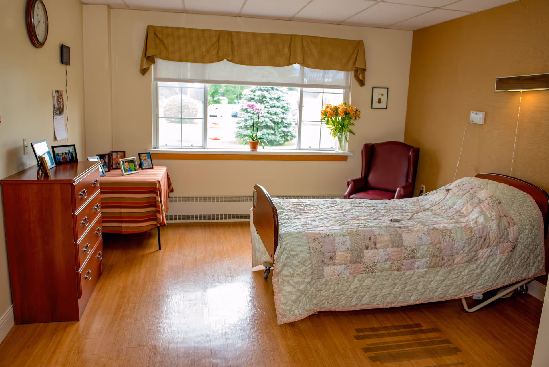 A cozy bedroom in a senior living facility featuring a bed, a red armchair, a dresser, and a window with flowers.