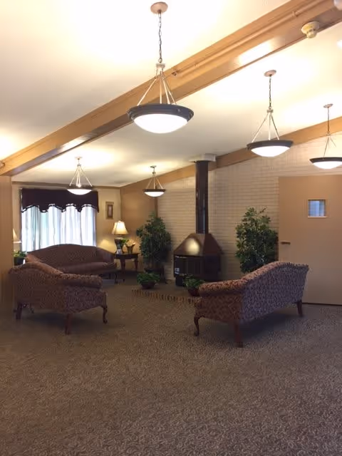 A cozy senior living facility common area with patterned upholstered sofas arranged around a small wood stove. The room has beige walls, carpeted floor, several hanging ceiling lights, potted plants, a side table with a lamp, and a window with curtains.