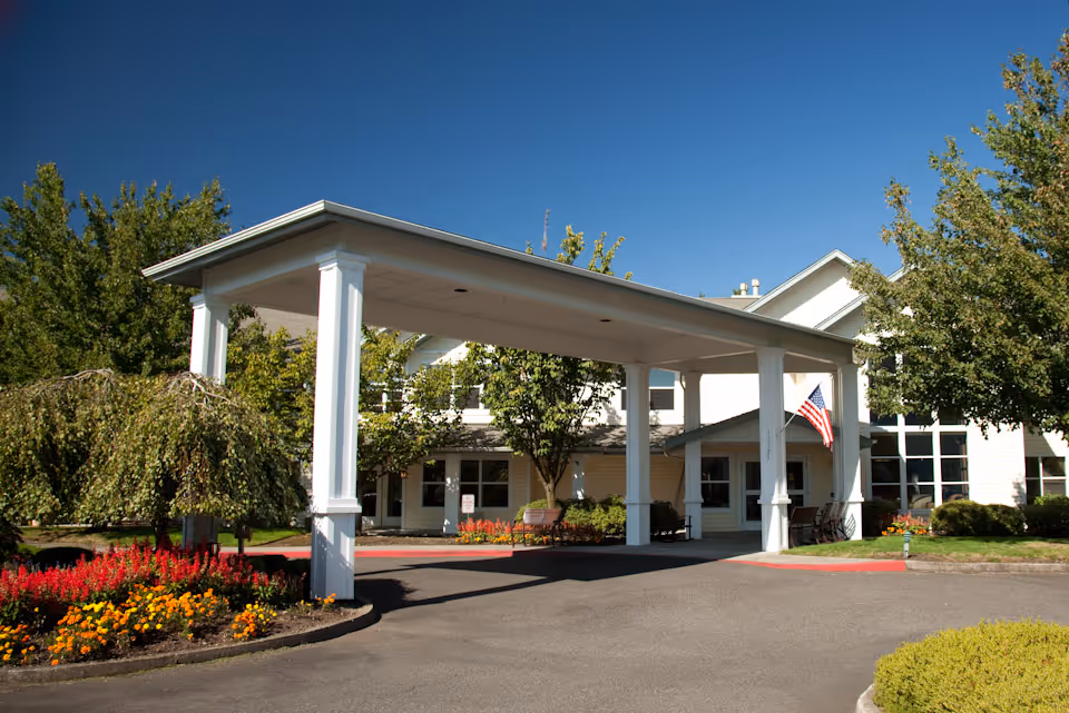 Front entrance of a white assisted living building with a covered porte-cochere, flowerbeds, and an American flag.
