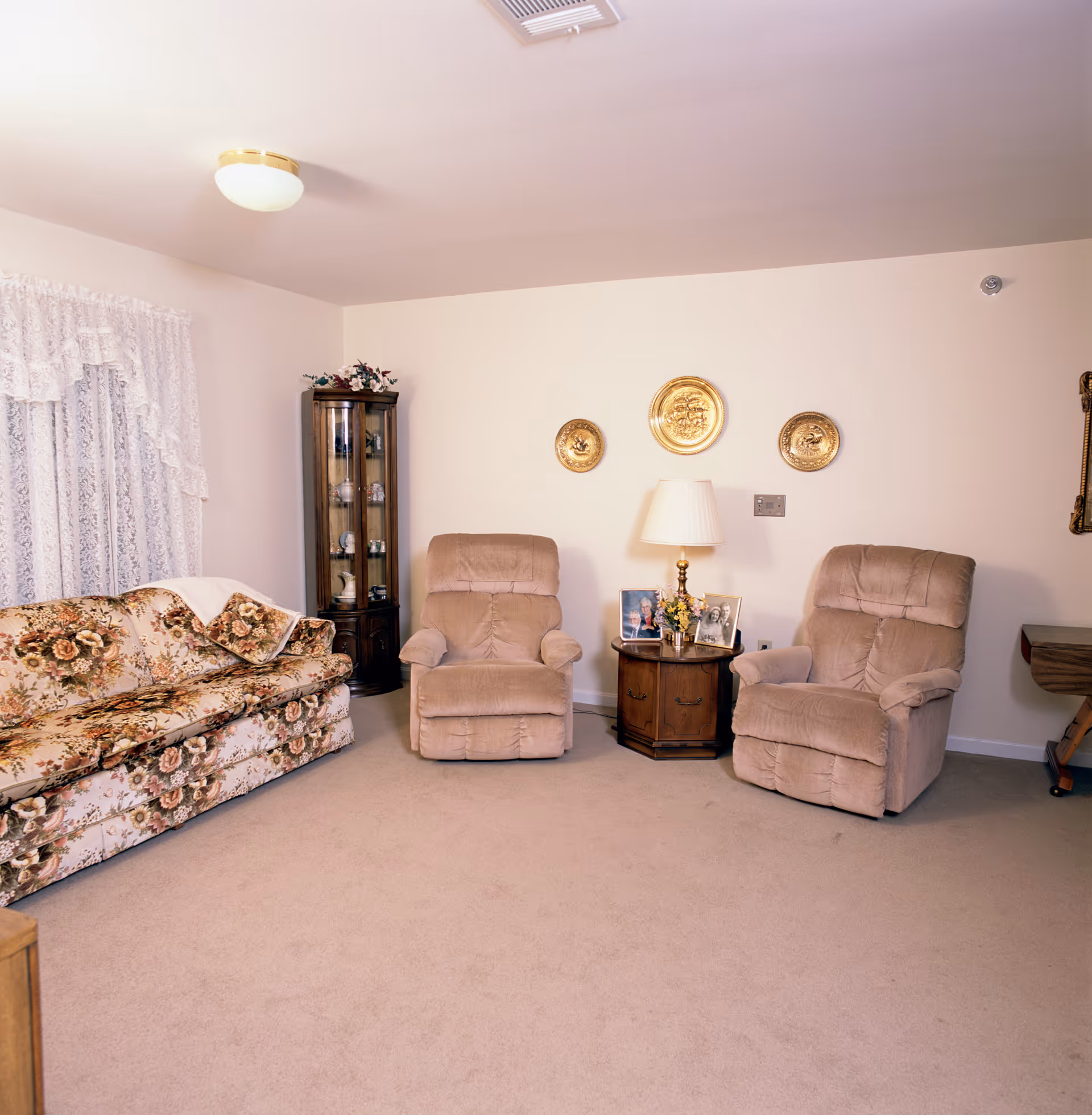 A cozy living room with a floral patterned sofa on the left and two beige recliner chairs facing forward. Between the chairs is a wooden side table with a lamp, framed photos, and a small flower arrangement. On the wall above the table are three decorative gold plates. A glass display cabinet with decorative items is in the corner near a window with lace curtains.
