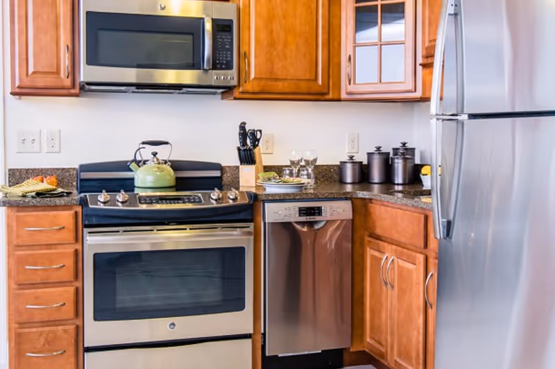 Modern kitchen with stainless steel appliances including a microwave, oven, dishwasher, and refrigerator. The kitchen has wooden cabinets, a granite countertop, a green kettle on the stove, a knife block, and some glassware and canisters on the counter.