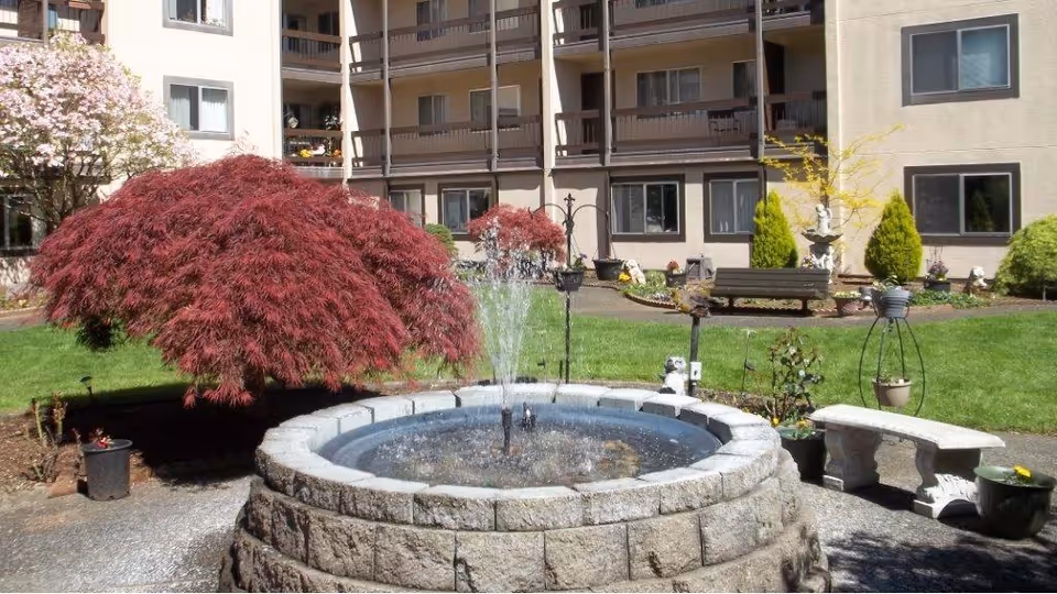 Stone fountain in a landscaped courtyard with a red-leafed tree, benches, potted plants, and a multi-story residential building in the background.