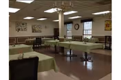 A dining room with multiple tables covered in light green tablecloths, each set with small flower vases. The room has brown and beige walls with framed artwork and windows with dark valances. The ceiling has fluorescent lighting and a chandelier.