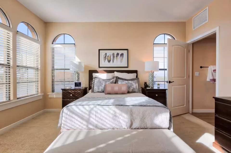 Sunlit bedroom with a neatly made bed centered between two nightstands, arched windows, and an open door to an adjacent room.