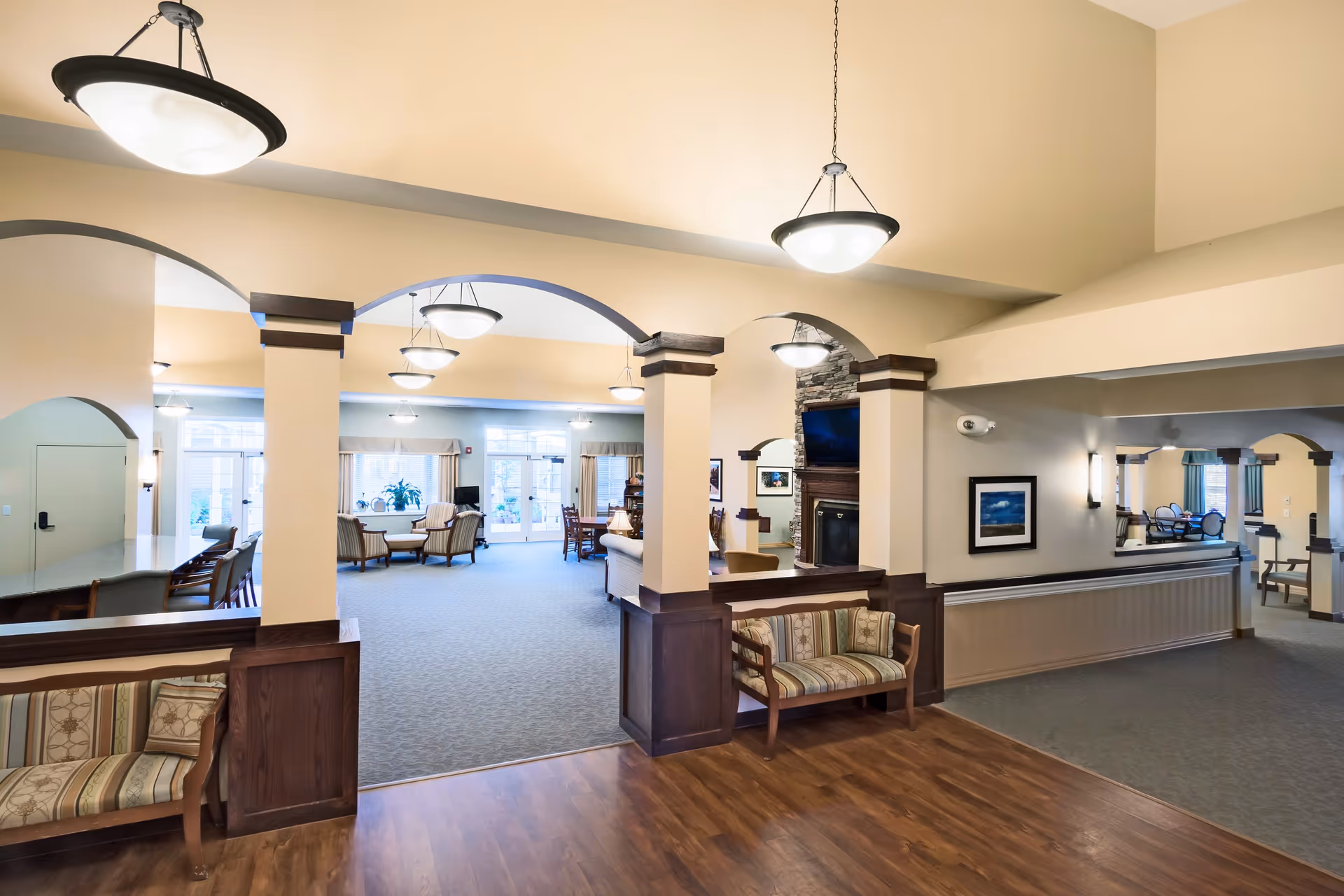 Spacious and well-lit common area in a senior living facility with arched openings, wooden benches with patterned cushions, hanging pendant lights, a stone fireplace with a mounted TV, and multiple seating arrangements including armchairs and tables near large windows.