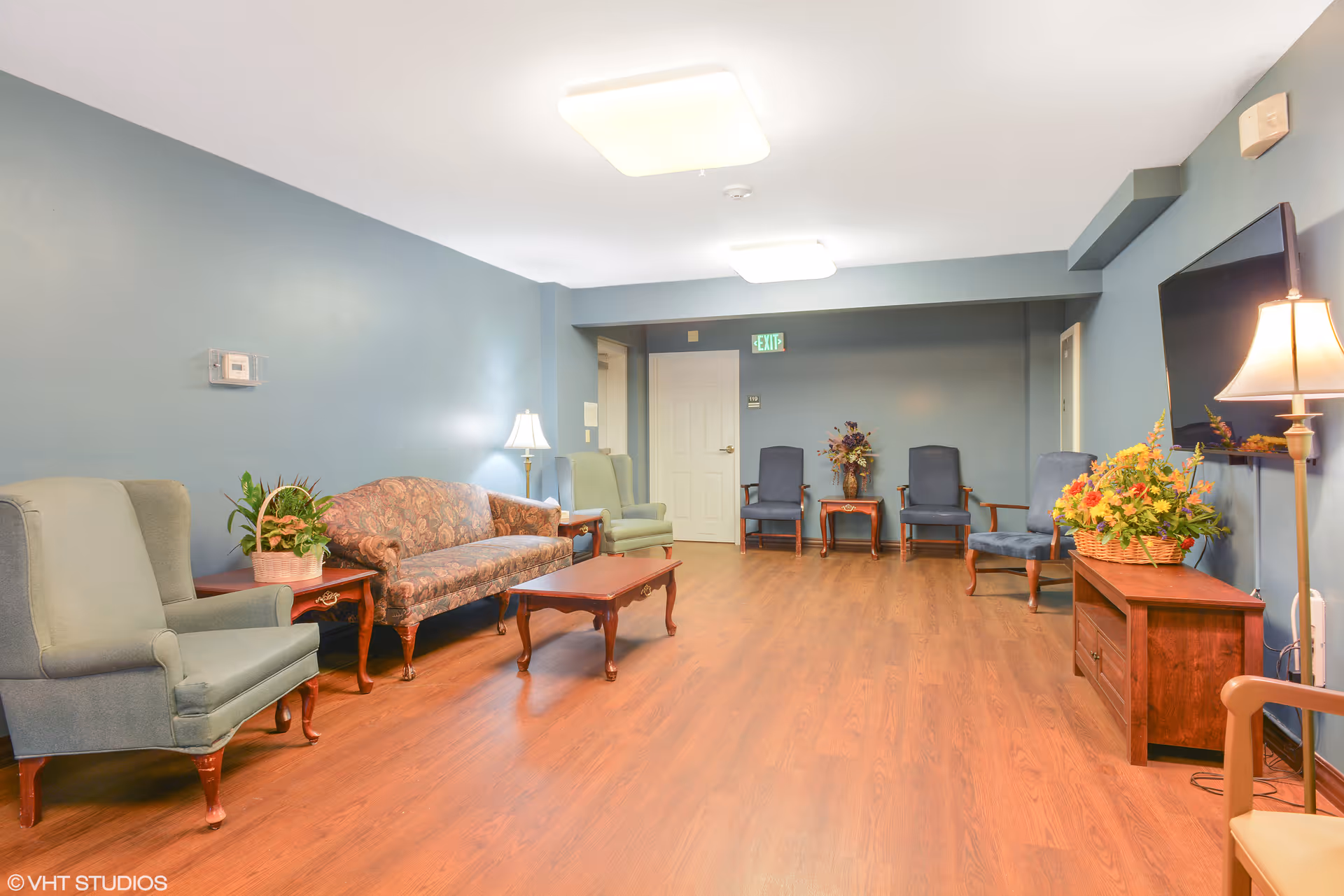 A spacious living room with wooden flooring and blue walls, furnished with various chairs, a patterned sofa, a wooden coffee table, side tables with lamps, and a TV mounted on the wall. There are decorative plants and flowers placed on the tables, and an exit sign above a white door in the background.