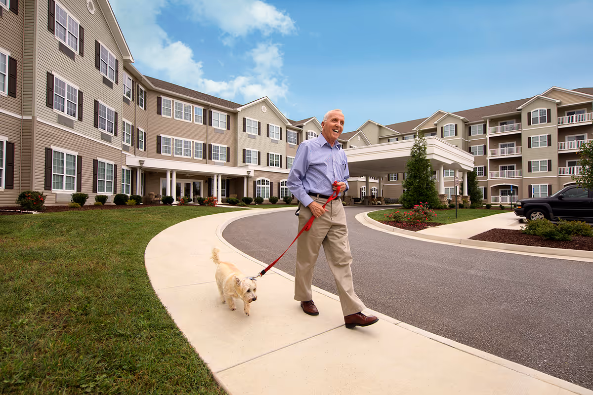 An older man walking a small dog on a sidewalk in front of a multi-story senior living building.