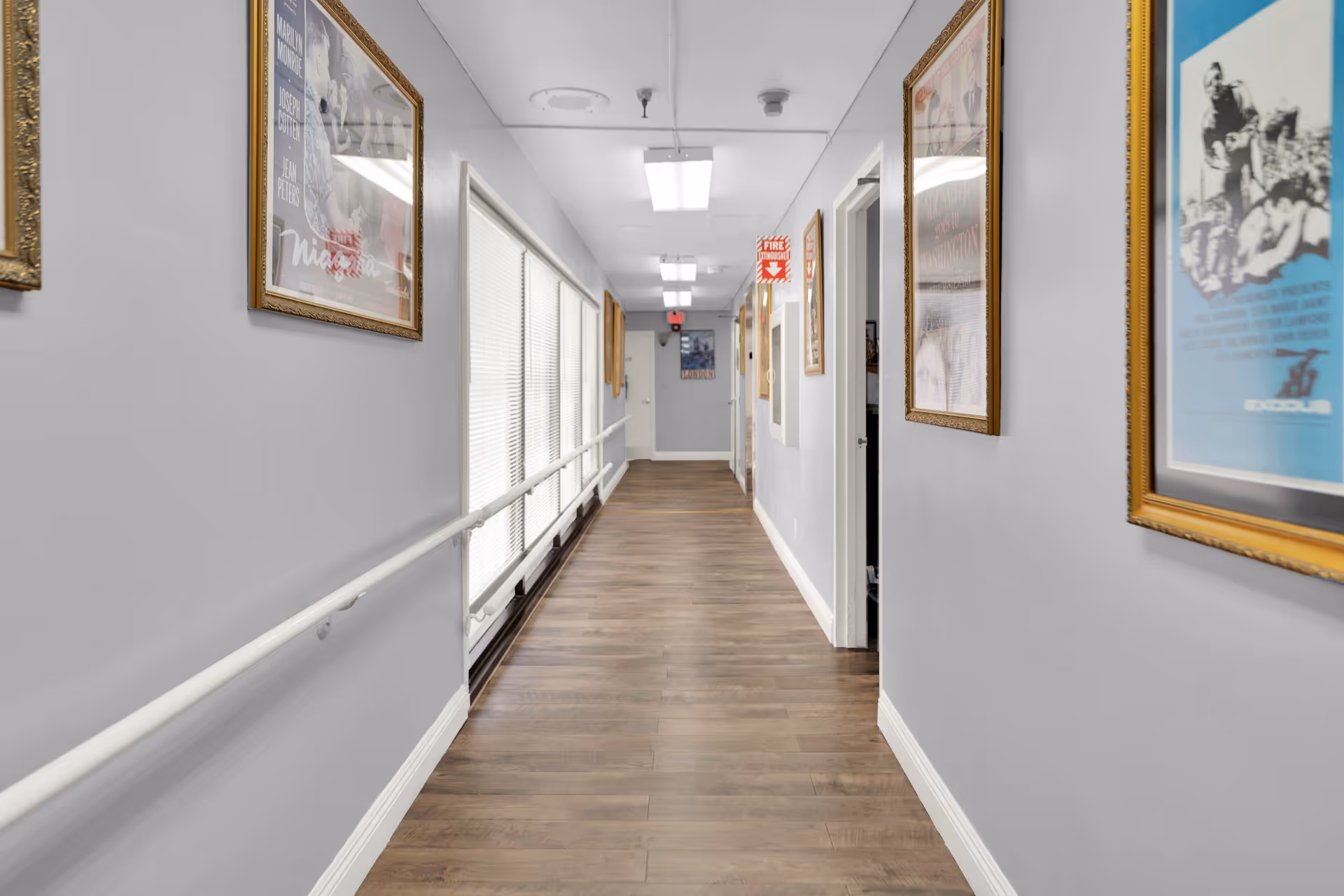 A bright interior hallway with wood flooring, wall-mounted handrails and framed artwork along the walls.