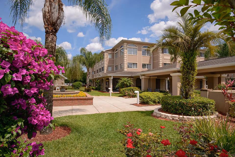 Exterior view of The Bridge at Orlando senior living facility showing a well-maintained garden with vibrant flowers, palm trees, and a paved walkway leading to a beige multi-story building under a partly cloudy blue sky.