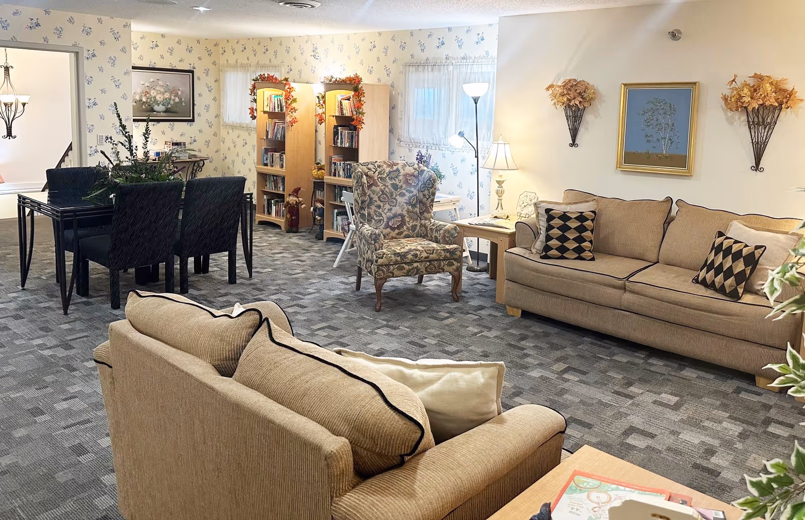 A cozy living room area in an assisted living facility featuring two beige sofas with patterned cushions, a floral armchair, a wooden side table with a lamp, and two wooden bookshelves decorated with autumn garlands. The room has patterned carpet flooring, floral wallpaper, and framed artwork on the walls. In the background, there is a dining area with a black table and chairs, and a chandelier hanging from the ceiling.