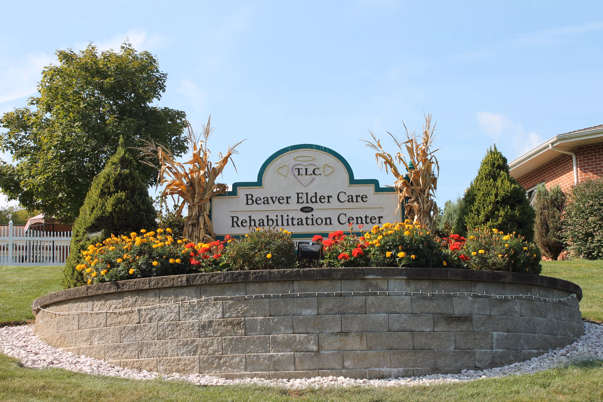 Outdoor view of a sign for Beaver Elder Care and Rehabilitation Center surrounded by a landscaped flower bed with yellow and red flowers, corn stalk decorations, and greenery. A brick building and trees are visible in the background under a clear blue sky.