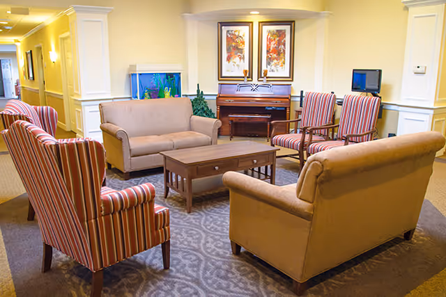 A cozy living room area with two beige sofas and three striped armchairs arranged around a wooden coffee table on a patterned rug. In the background, there is a piano with two framed artworks above it, a fish tank, and a small TV on a stand. The walls are painted light yellow with white trim and columns.