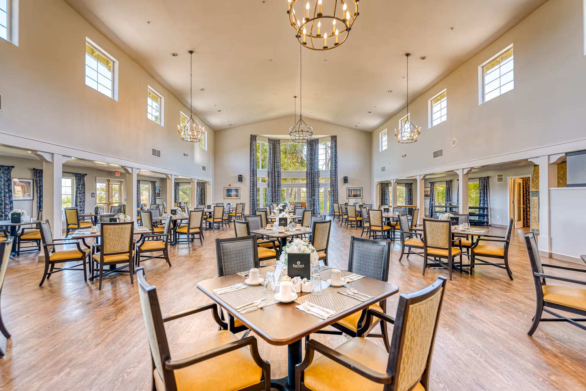 A spacious and well-lit dining room with multiple tables and chairs arranged neatly. The room features high ceilings with hanging chandeliers, large windows with blue patterned curtains, and wooden flooring. Each table is set with placemats, napkins, cups, and a small floral centerpiece.