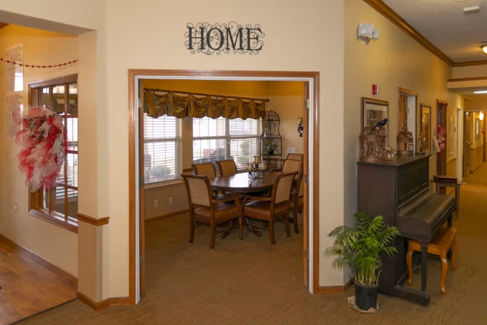 Entrance from a hallway into a dining room with a round table and chairs, a piano and decorative plants under a "HOME" sign.