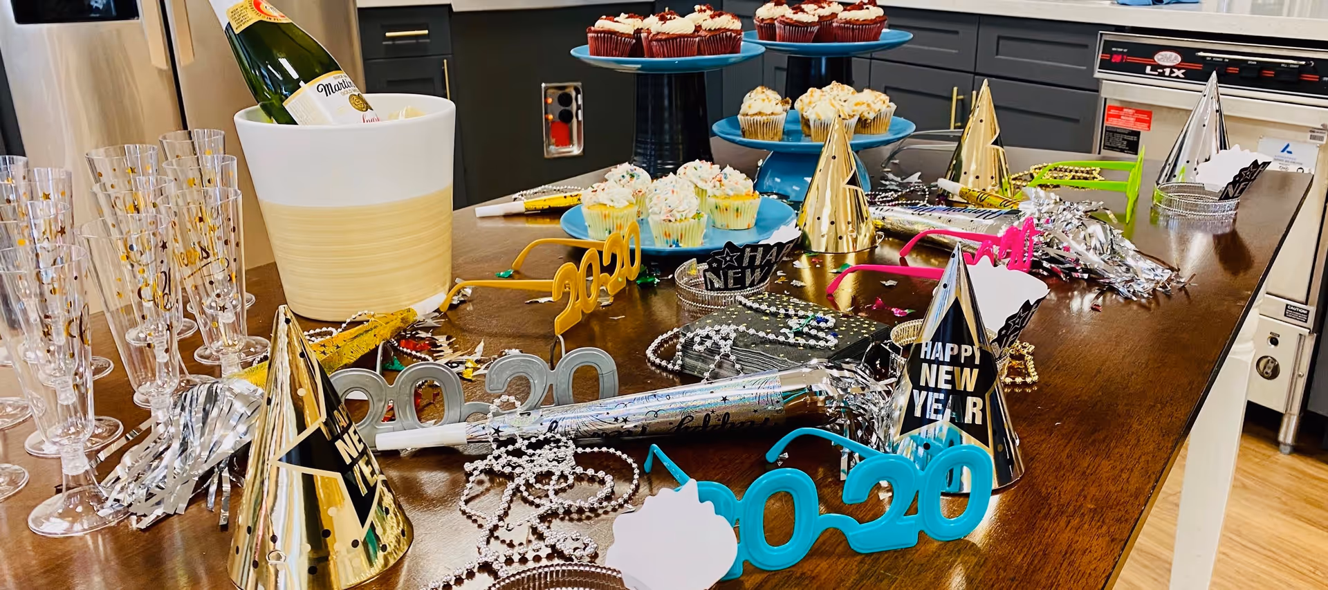 Kitchen island arranged for a New Year's celebration with champagne in an ice bucket, plastic champagne flutes, party hats, '2020' glasses, cupcakes and noisemakers.