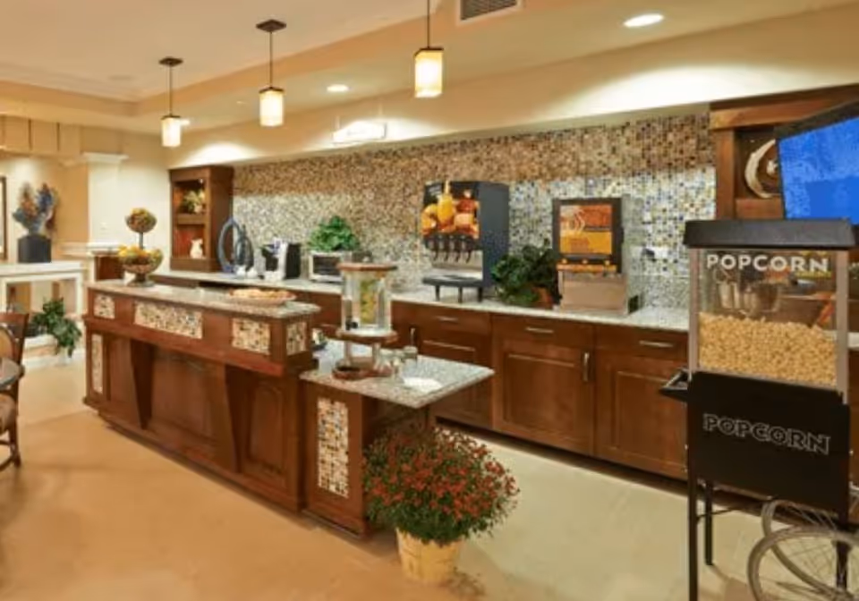 Interior view of a refreshment area in an assisted living facility featuring a wooden counter with mosaic tile accents, a popcorn machine, beverage dispensers, a microwave, and decorative plants under warm lighting.
