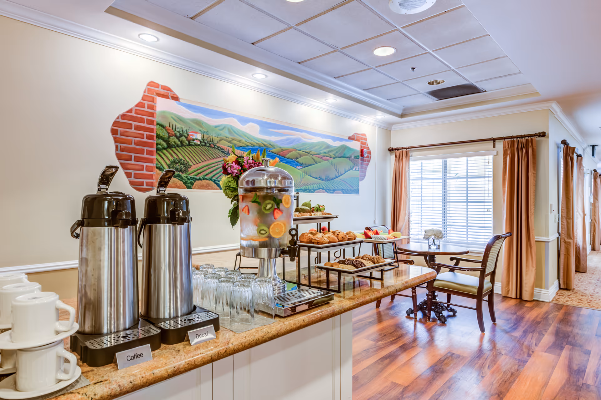 A refreshment area in a senior living facility with two large coffee dispensers labeled Coffee and Decaf, a glass beverage dispenser filled with water and fruit slices, and trays of assorted pastries and muffins. The area has a countertop with stacked white cups and glasses, a colorful mural of a countryside landscape on the wall, and a small table with chairs near a window with brown curtains. The floor is wooden, and the ceiling has recessed lighting.