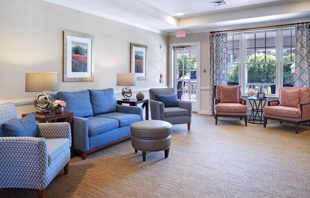 A bright and cozy living room with a blue sofa, two patterned armchairs, two coral armchairs, a round ottoman, two side tables with lamps, framed artwork on the wall, large windows with patterned curtains, and a glass door leading outside.