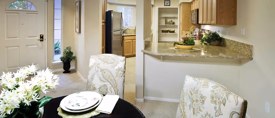 View of a dining area with a round table set with plates and napkins, floral upholstered chairs, and a vase of white flowers. In the background, there is a kitchen with wooden cabinets, a granite countertop, and a stainless steel refrigerator. To the left, there is a white front door with a window and a potted plant nearby.